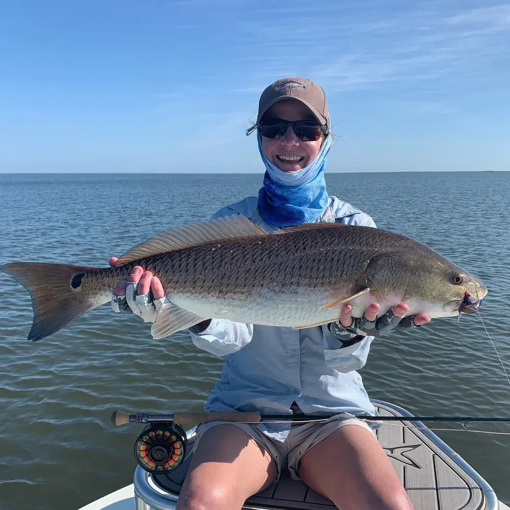 Person on a boat holding a large redfish; sunny day, water in the background.