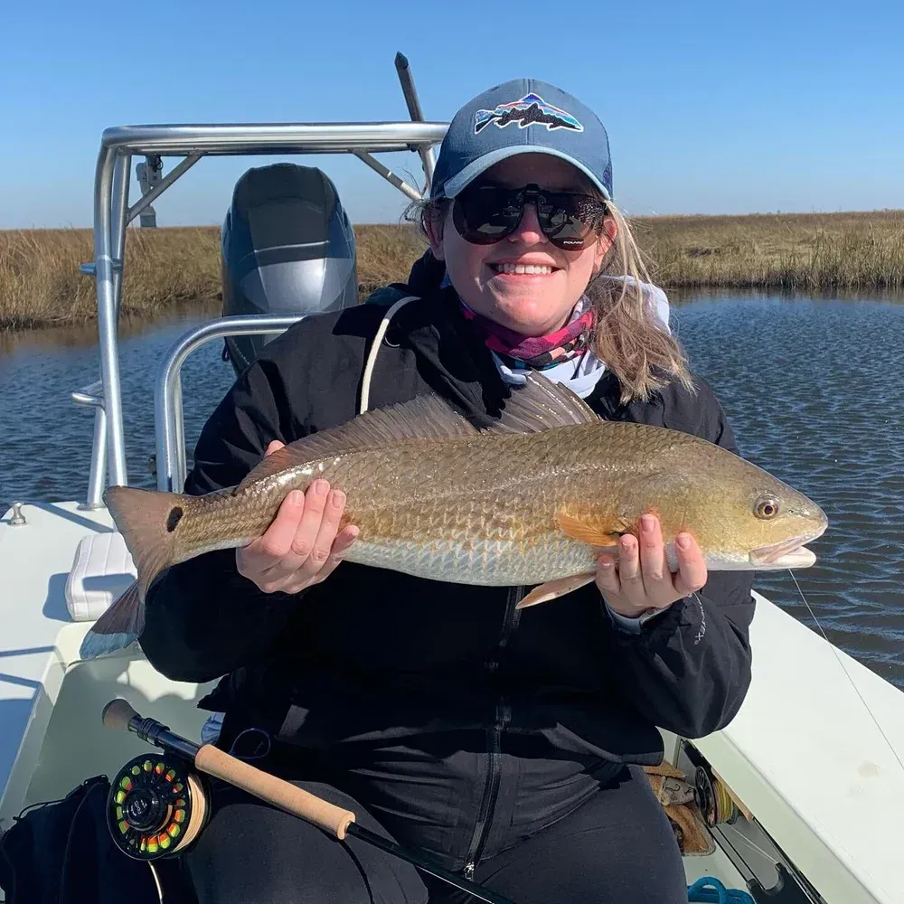 Woman smiles, holding a redfish on a boat in marsh. She wears a hat and sunglasses.