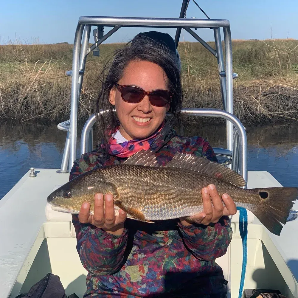 Woman smiling, holding a redfish on a boat in a marsh. Sunny day, wearing sunglasses and patterned jacket.
