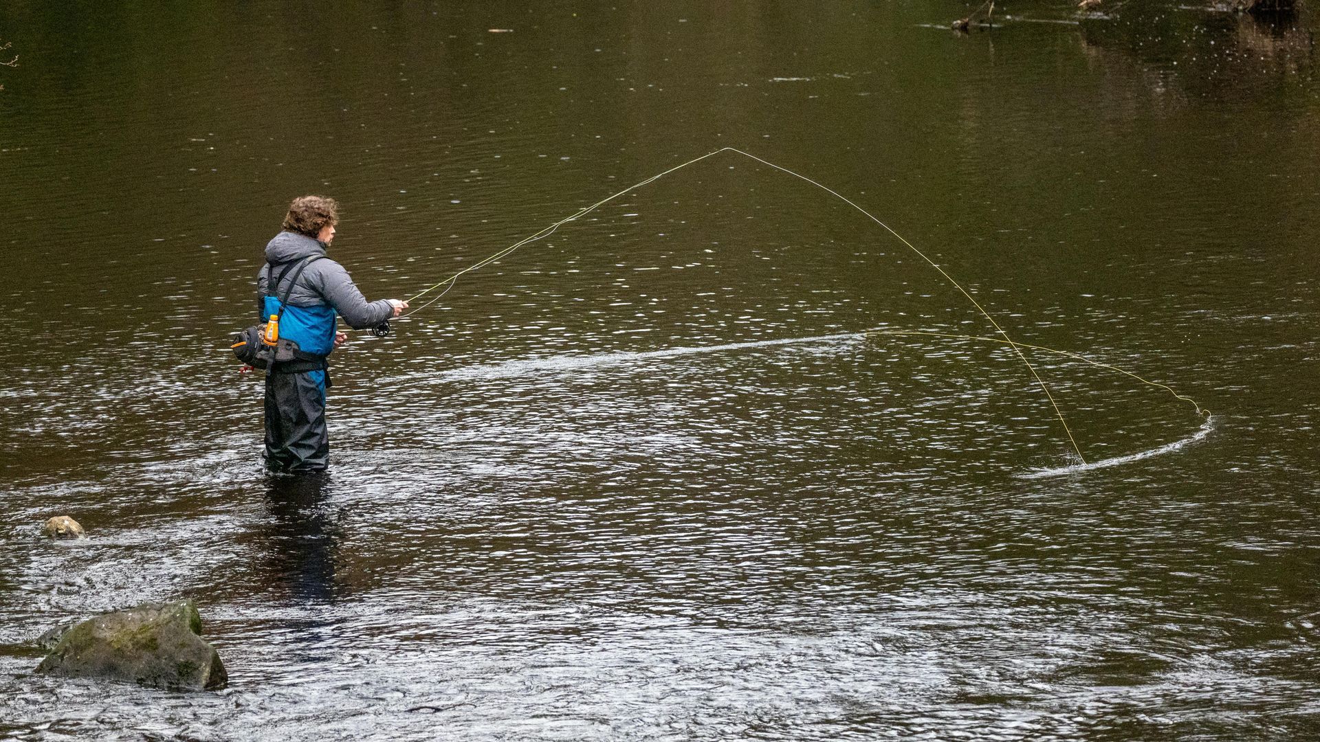 Person fly-fishing in a river, casting a line with ripples around them.