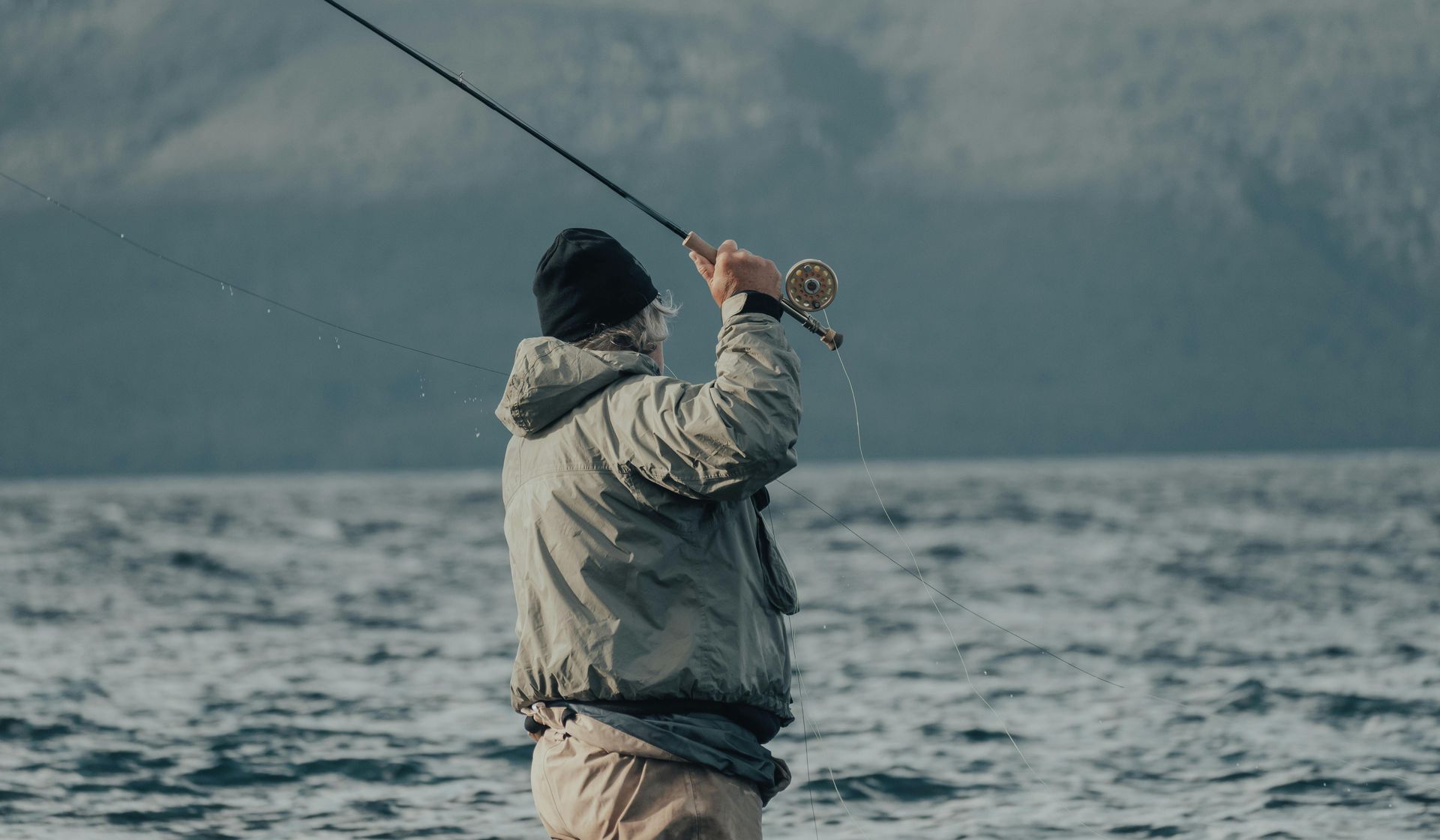 Person fishing with a fly rod in a body of water, wearing a jacket and a beanie. Mountains in the background.