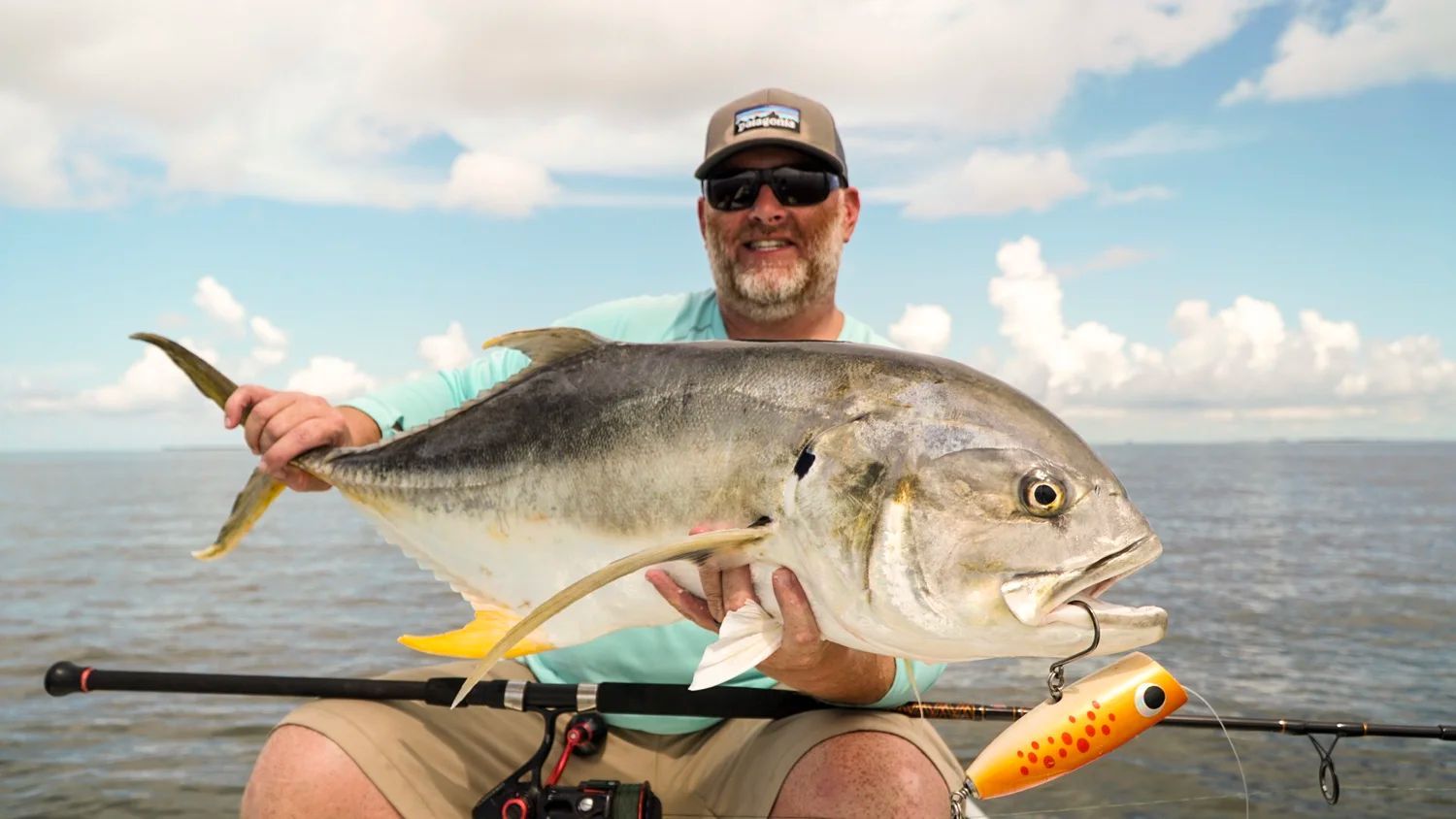 Man on a boat holds a large, silver fish with a lure in its mouth; blue sky in the background.