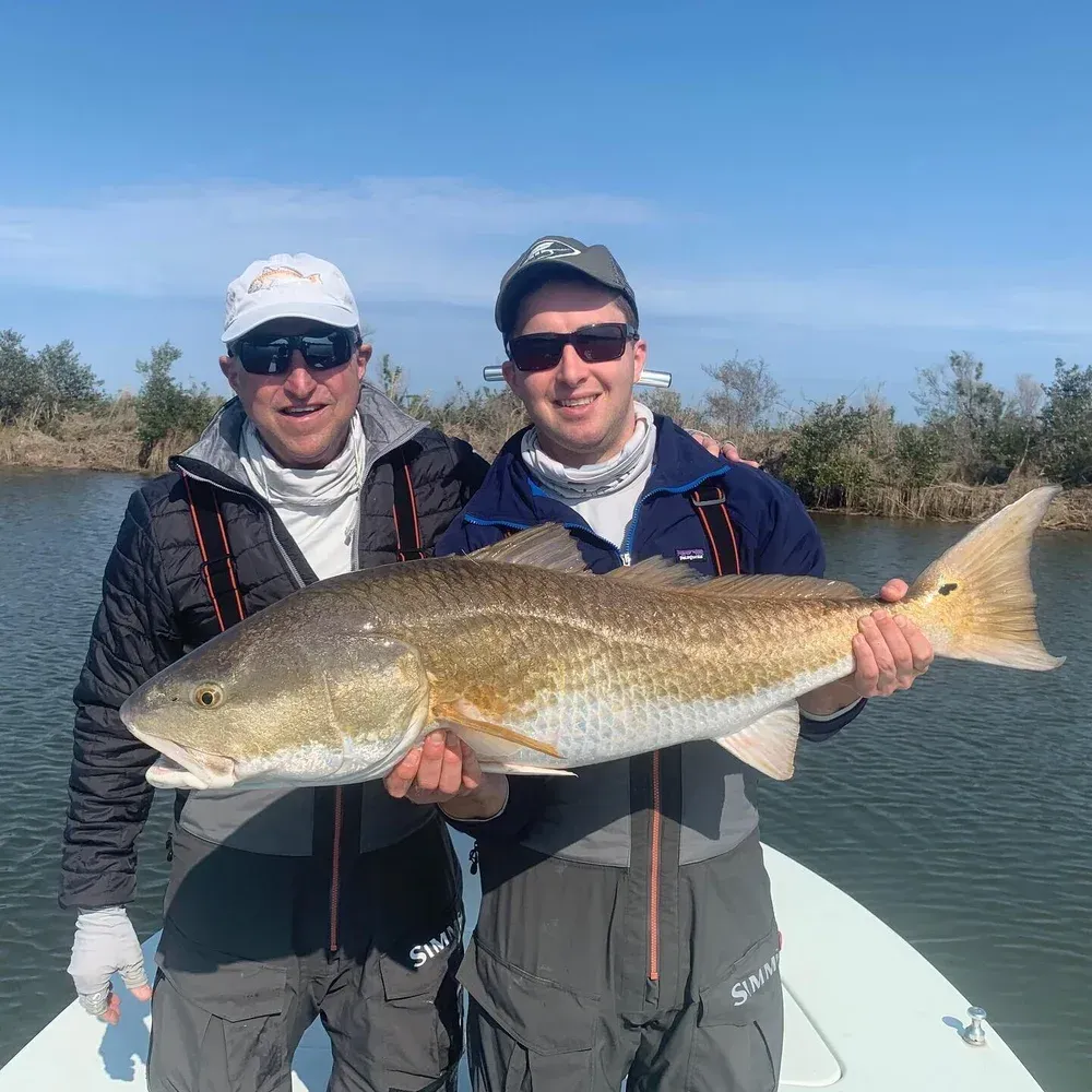 Two men on a boat hold up a large, reddish-brown fish. They are smiling, with greenery in the background.