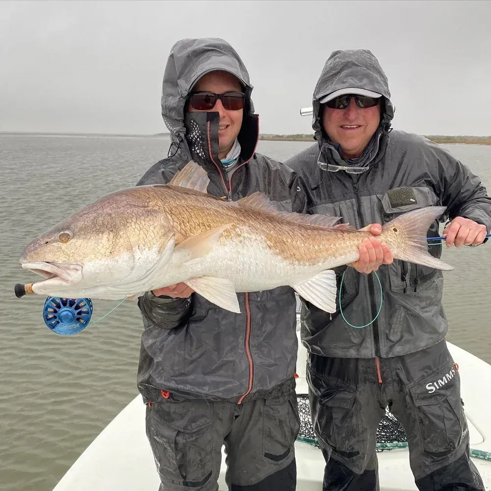 Two men on a boat holding a large redfish, both wearing rain gear and smiling.