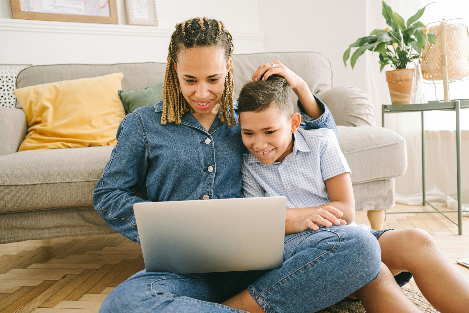 Woman and boy sit on floor looking at laptop, woman's hand on boy's head. Living room setting.