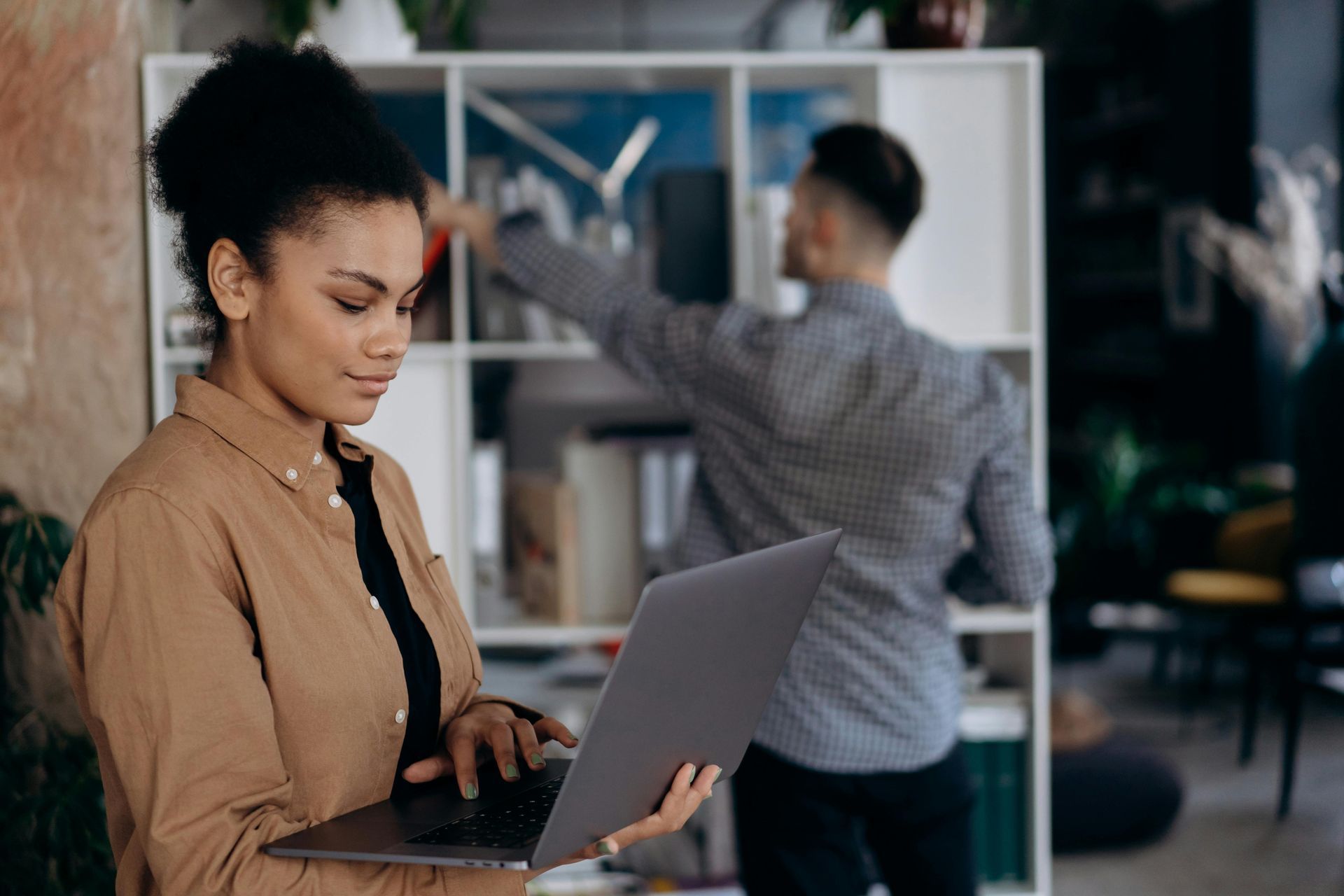 Woman with laptop, looking focused; man reaching for items on bookshelf in background.