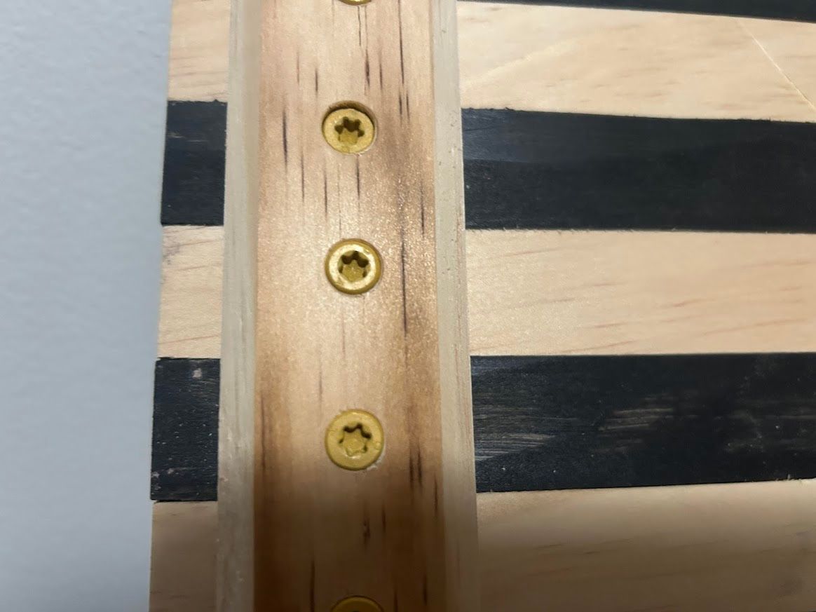 Close-up of wooden frame with black and tan stripes, secured with gold screws.