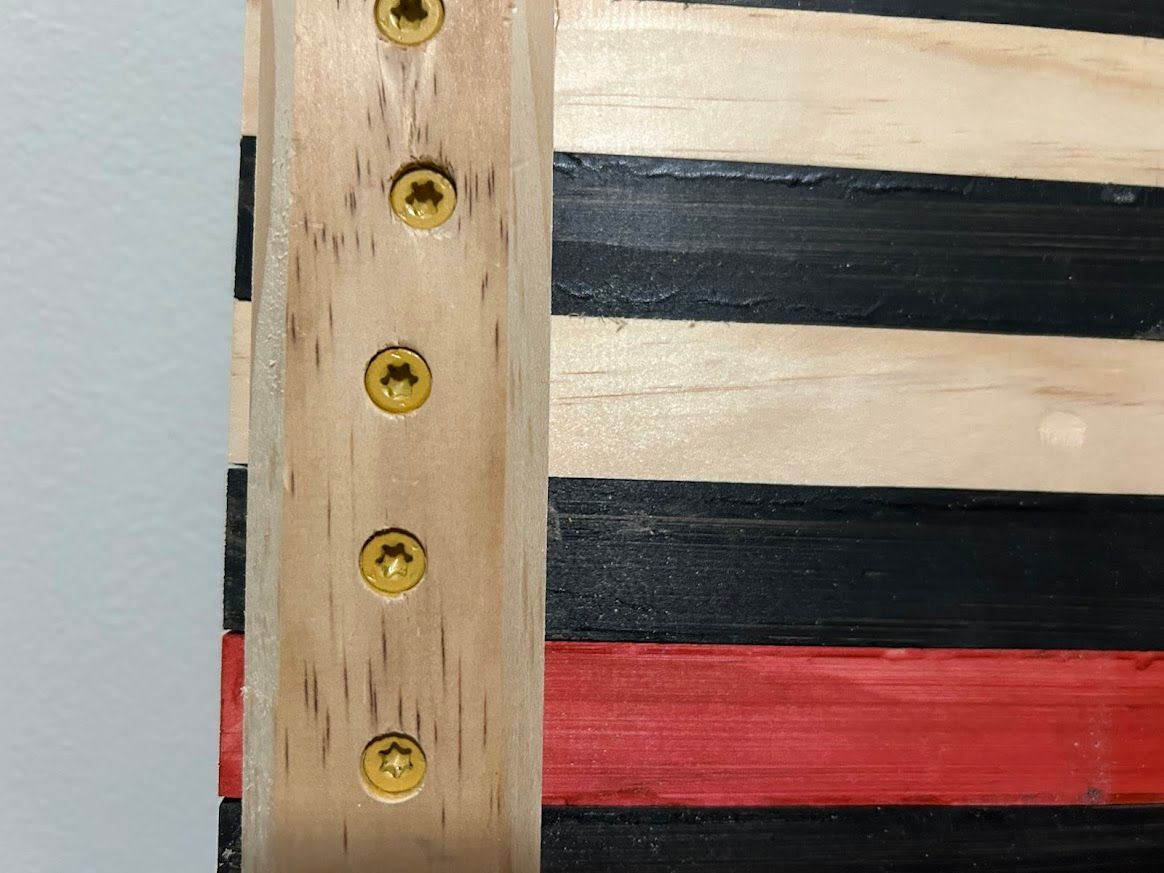 Wooden flag with black, red, and beige stripes, held together with gold screws.
