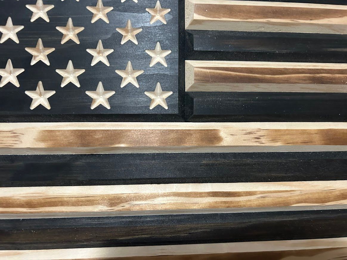 Wooden American flag with carved stars, black and tan stripes, on a wooden surface.