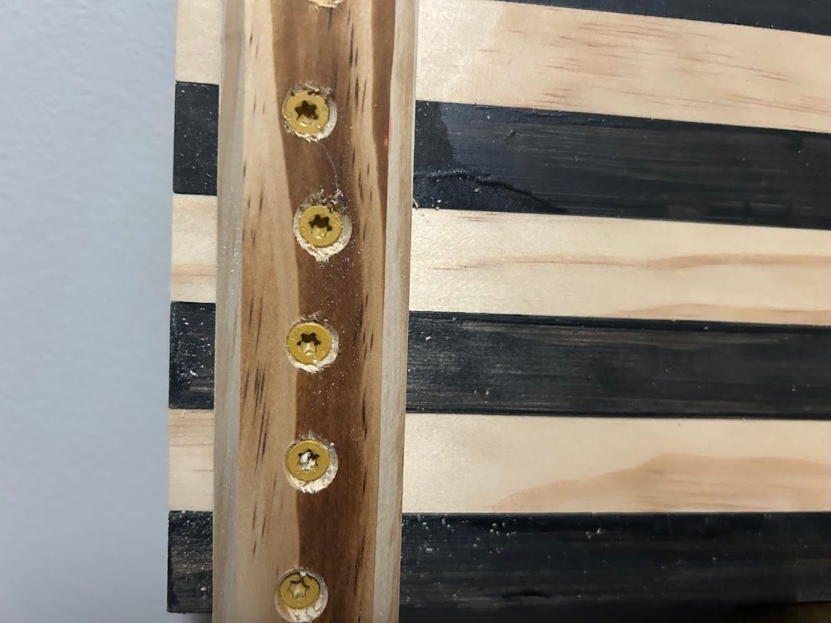 Close-up of a wooden American flag, showing the screw attachment of a vertical piece to the black and natural wood stripes.