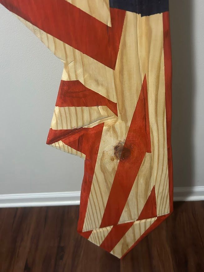 Wooden American flag art with red and white stripes, blue corner, displayed against a gray wall and brown floor.