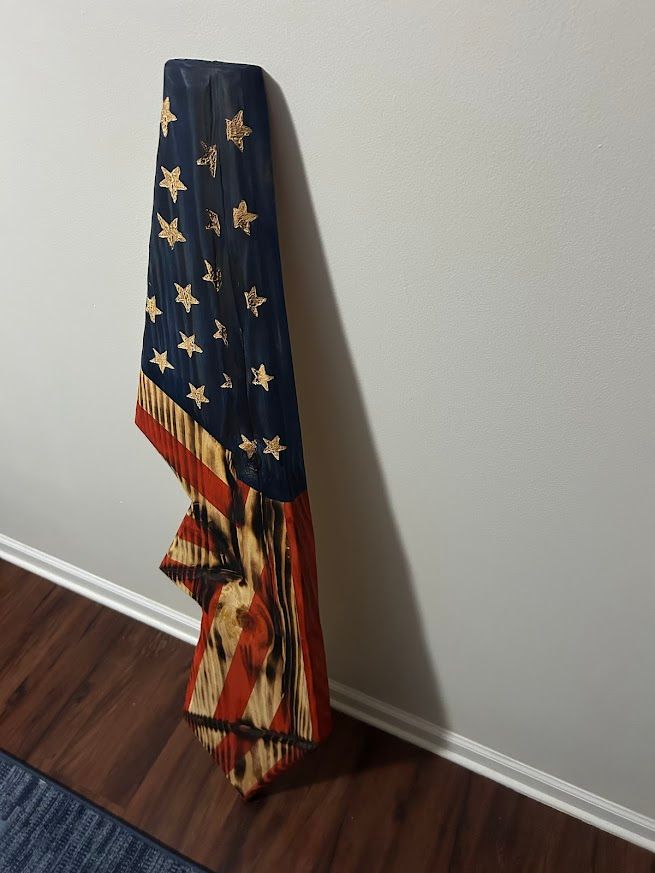 Wooden American flag sculpture leaning against a white wall and wood floor.