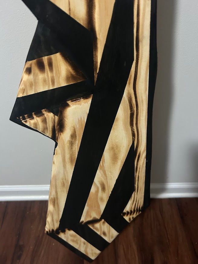 Wooden art: wood-burned with black stripes. Beige wood shows through, leaning against a gray wall, on a wood floor.