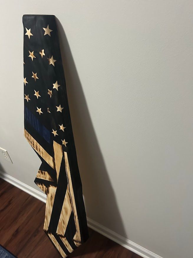 Burnt wood American flag with blue stripe. Leaning against a white wall and wood floor.