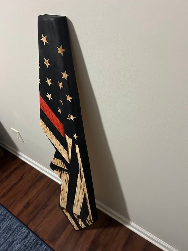 Wooden American flag with a red stripe, leaning against a white wall and brown floor.