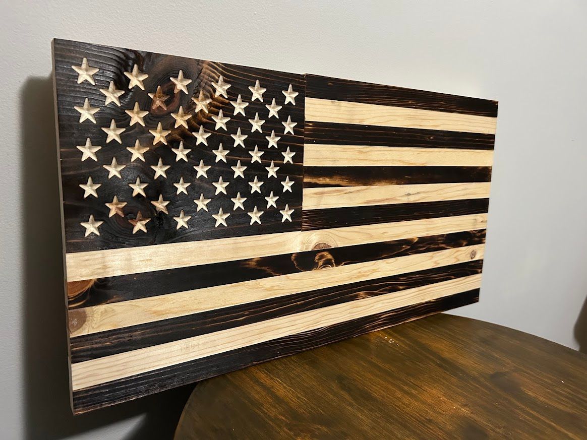 Wooden American flag with burnt stripes and star field, on a dark wooden table.