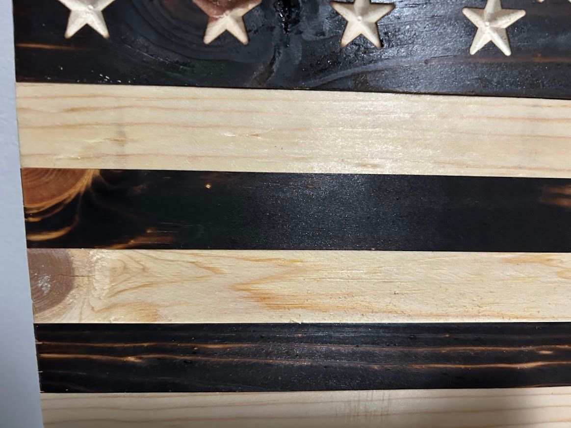Wooden American flag, with alternating burnt black and natural wood stripes, and carved white stars in a blue field.