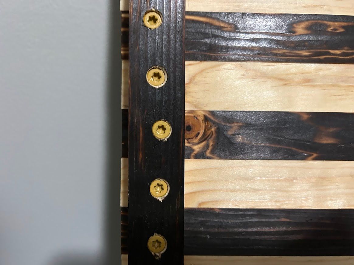 Close-up of a wooden American flag with black and tan stripes, held together with screws and a dark wooden border.