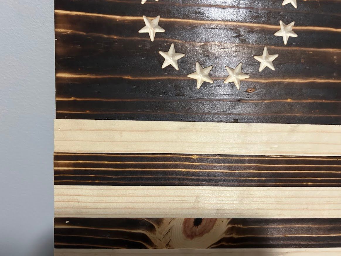 Wooden flag with burned stripes, carved stars in a circle, on a wall.