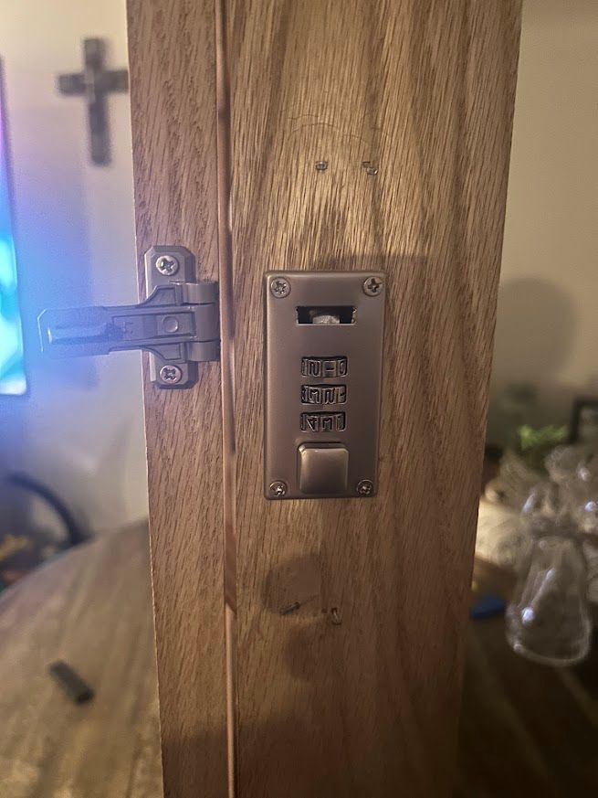 Wooden door with a combination lock and hinge. The numbers on the lock are 285. A cross is in the background.