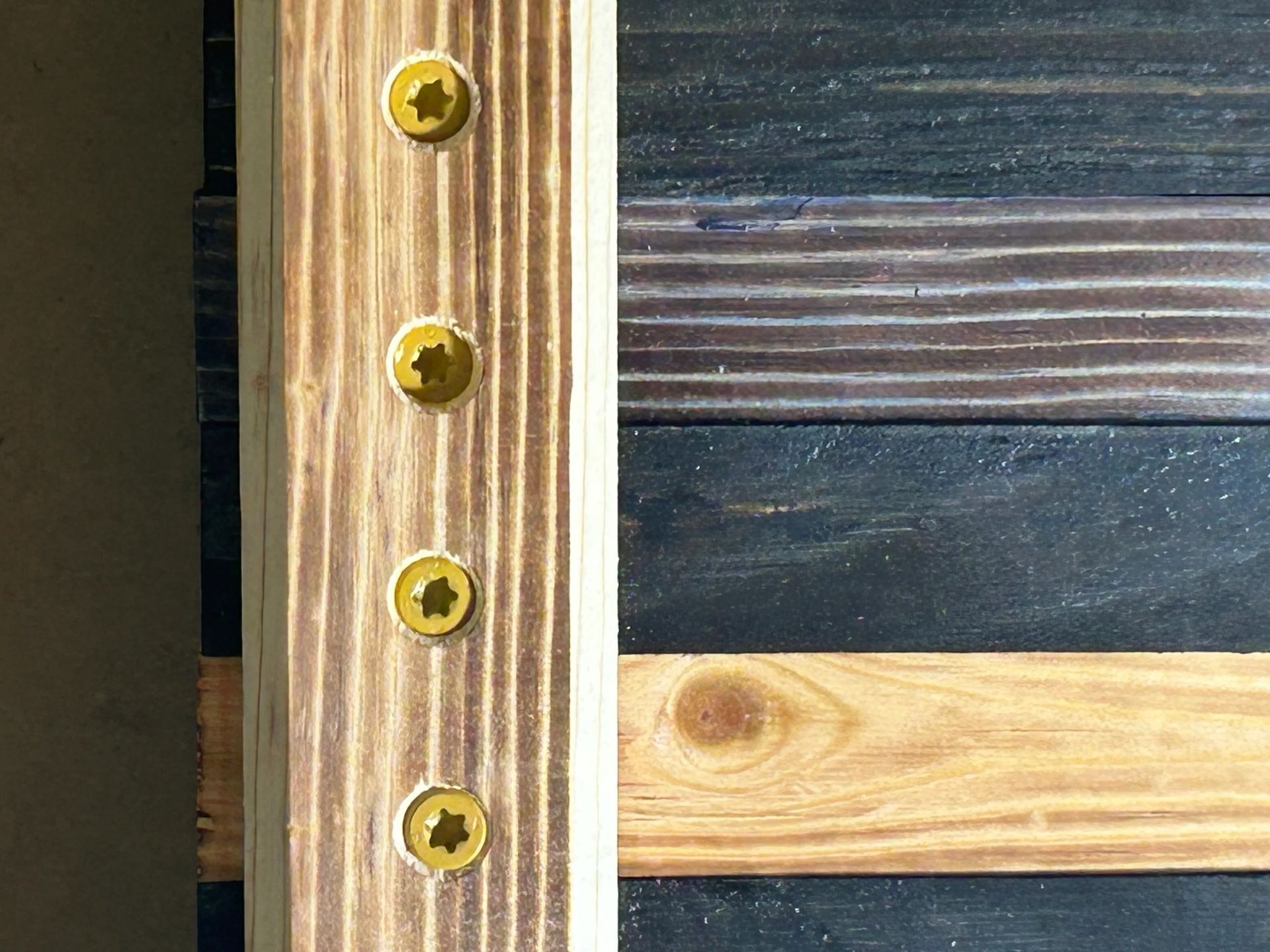 Wooden planks joined by a vertical board with four gold screws; the planks are stained black and natural wood.