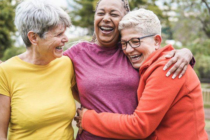 Three older women are hugging each other and laughing in a park.