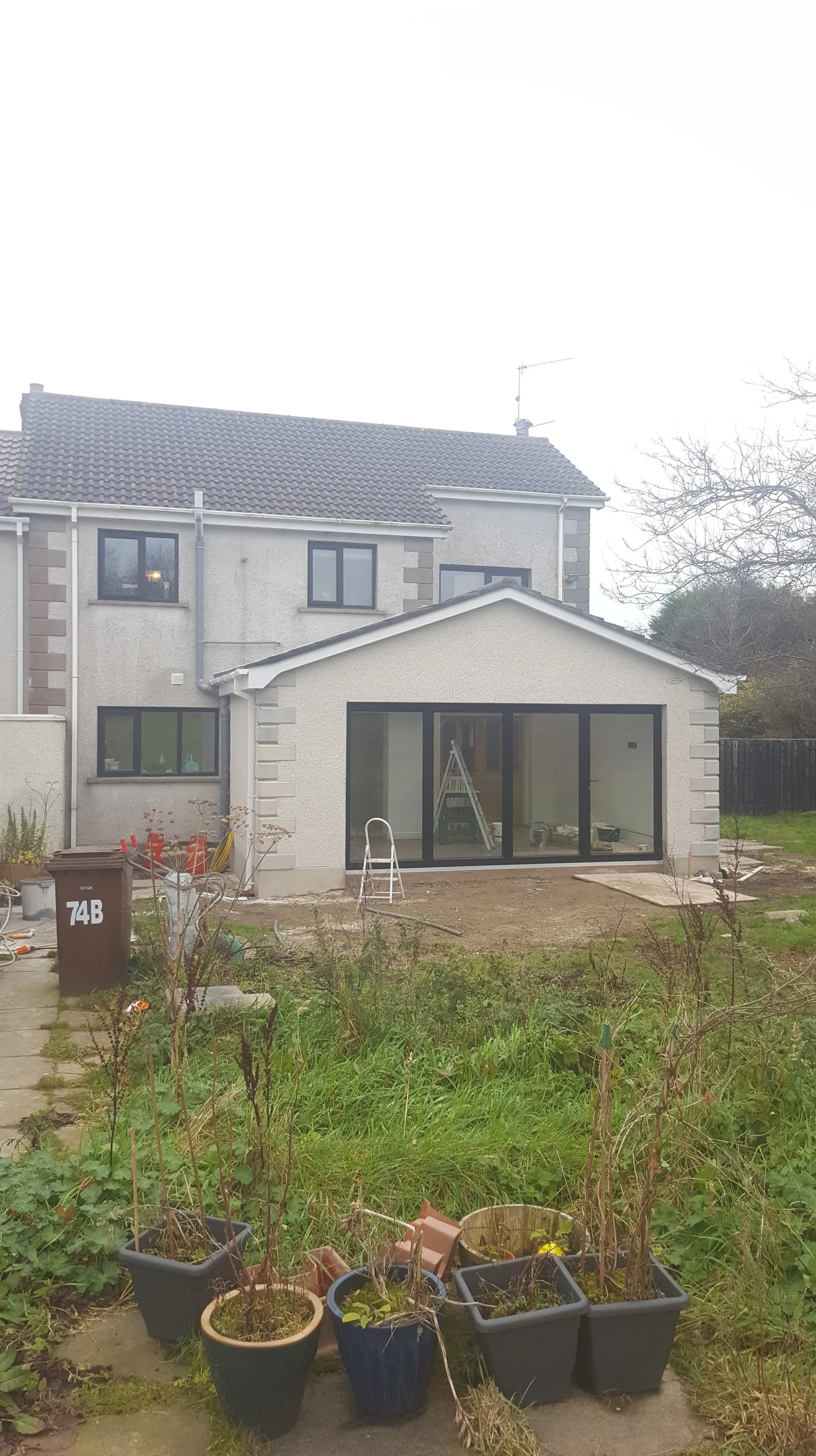 Backyard view of a two-story house with a newly constructed extension featuring black framed sliding doors. Overcast day.