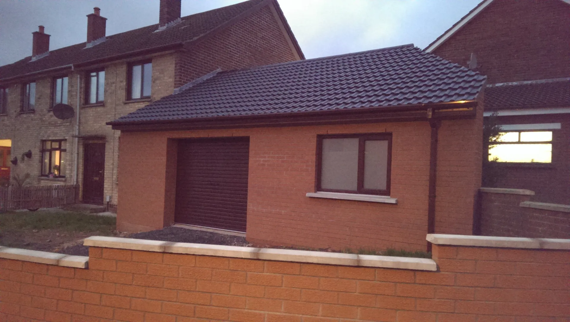 A brick building with a dark roof and a closed garage door and window. Behind it, a row of houses.