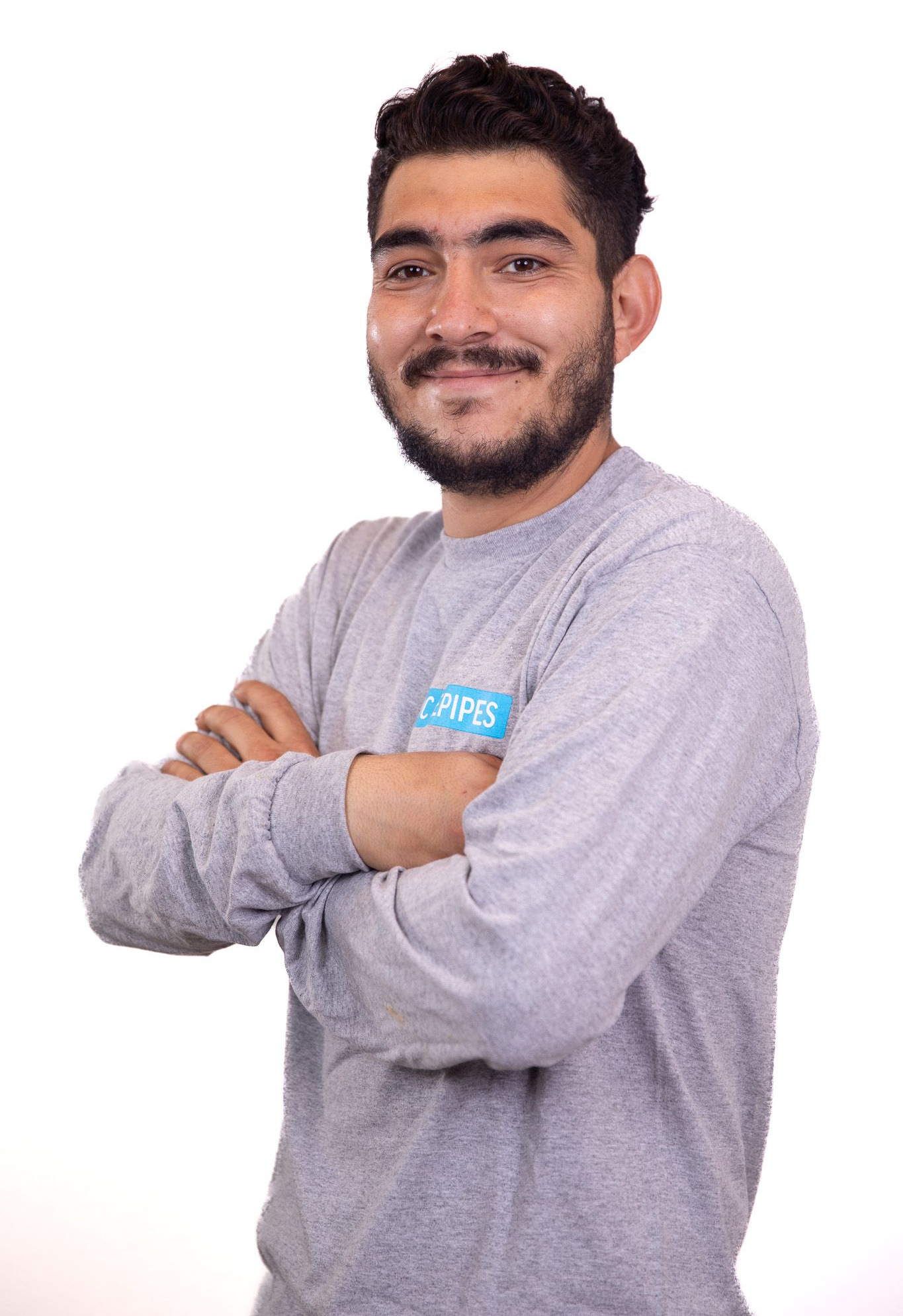 Man with beard, arms crossed, smiling. Wearing grey long sleeve shirt with blue logo.