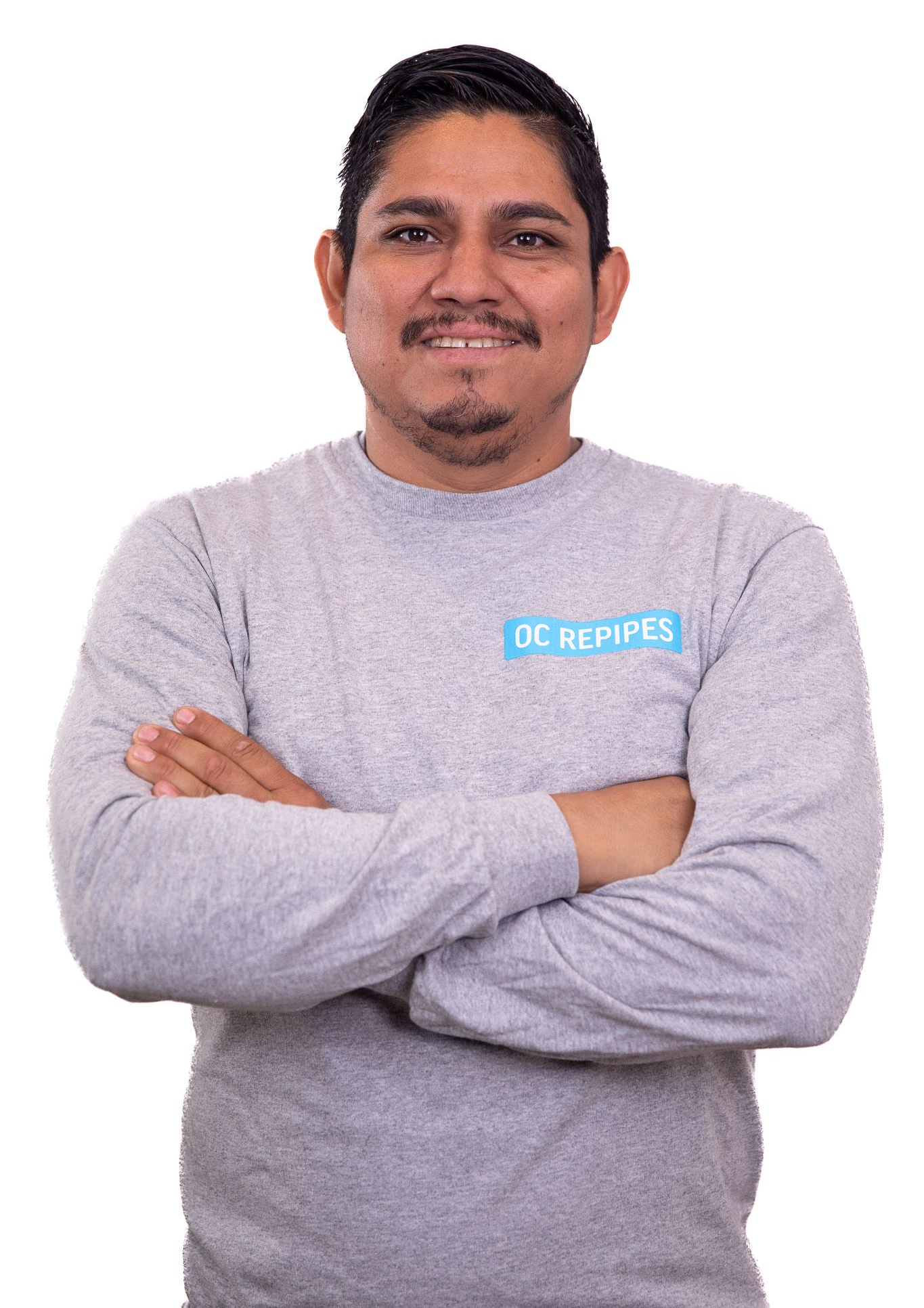 Man with arms crossed, wearing a gray shirt with a logo. He smiles at the camera, against a white backdrop.