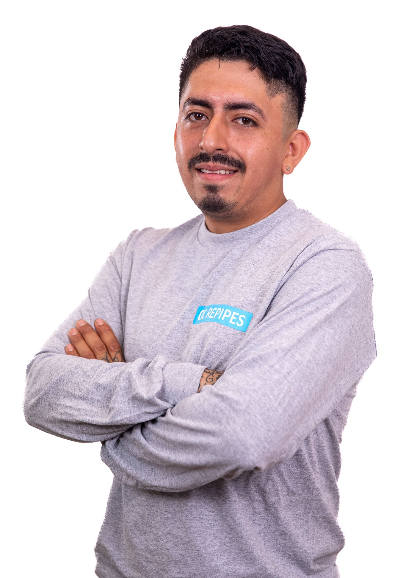Man with arms crossed, wearing gray long-sleeved shirt with logo, smiling.