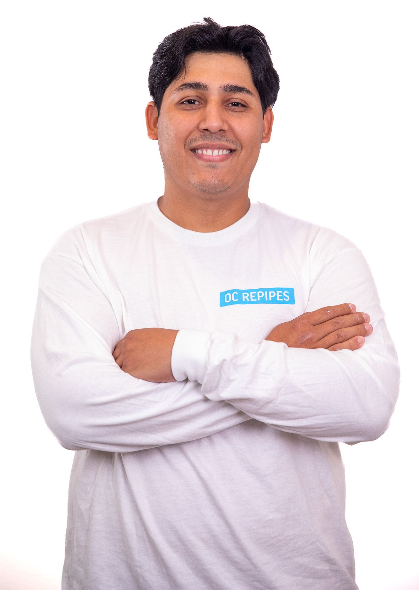 Man in white long-sleeve shirt with arms crossed, smiling at the camera. Text on shirt reads 