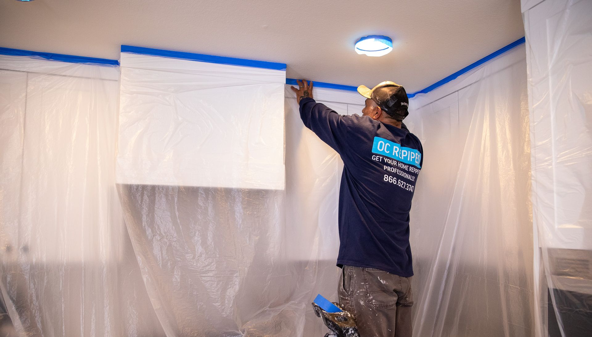 Two plumbers re-piping a ceiling. One wears a hat with a headlamp and a shirt that says