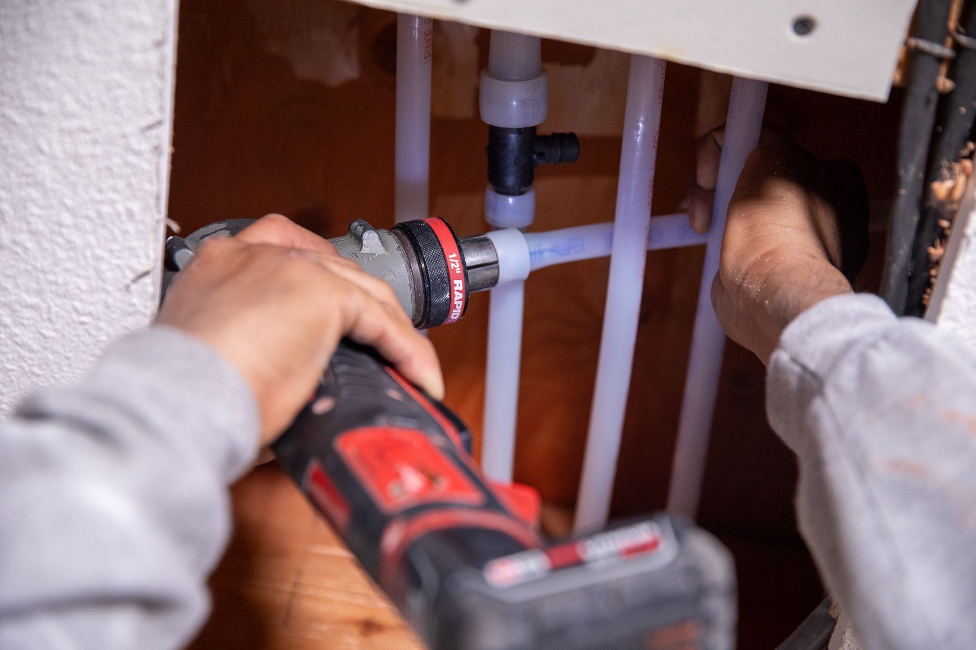 A person using a power tool to work on PEX plumbing pipes inside a wall.