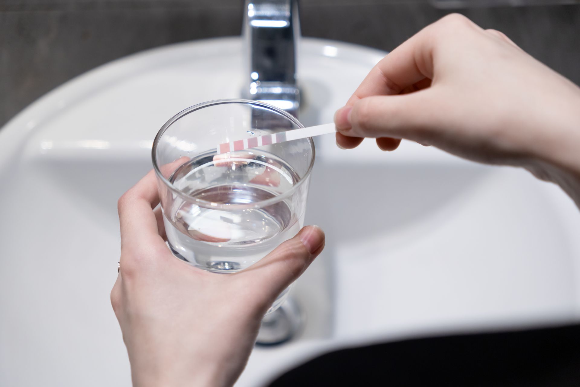 Person testing water in a glass with a test strip, next to a white sink.