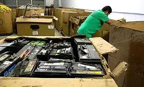 A person in a green shirt sorts used lead-acid batteries into a large cardboard box in a warehouse setting.