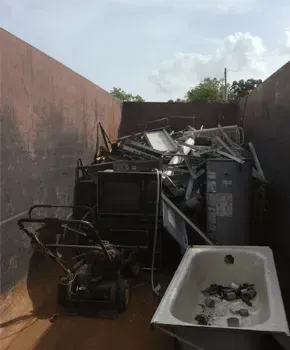 A large metal dumpster filled with scrap metal, including a lawn mower, a bathtub, and discarded electronic appliances.
