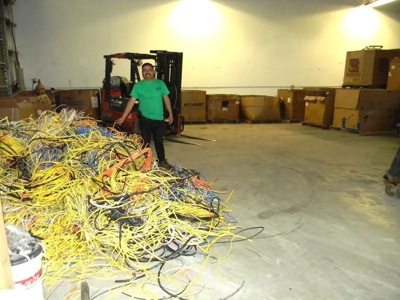 A person in a green shirt stands in a warehouse next to a large pile of yellow and black scrap cables and a forklift.