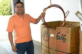 A person in an orange polo shirt stands next to a large cardboard box filled with coils of bare copper wire.