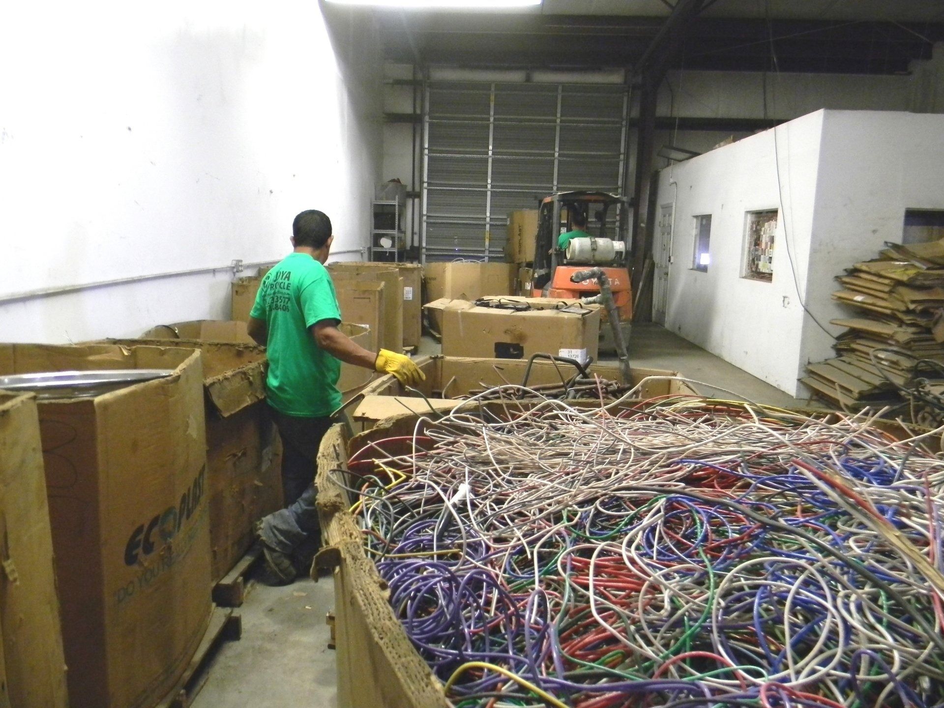 A worker in a green shirt sorting through a large bin of colorful, tangled scrap wires in a warehouse.