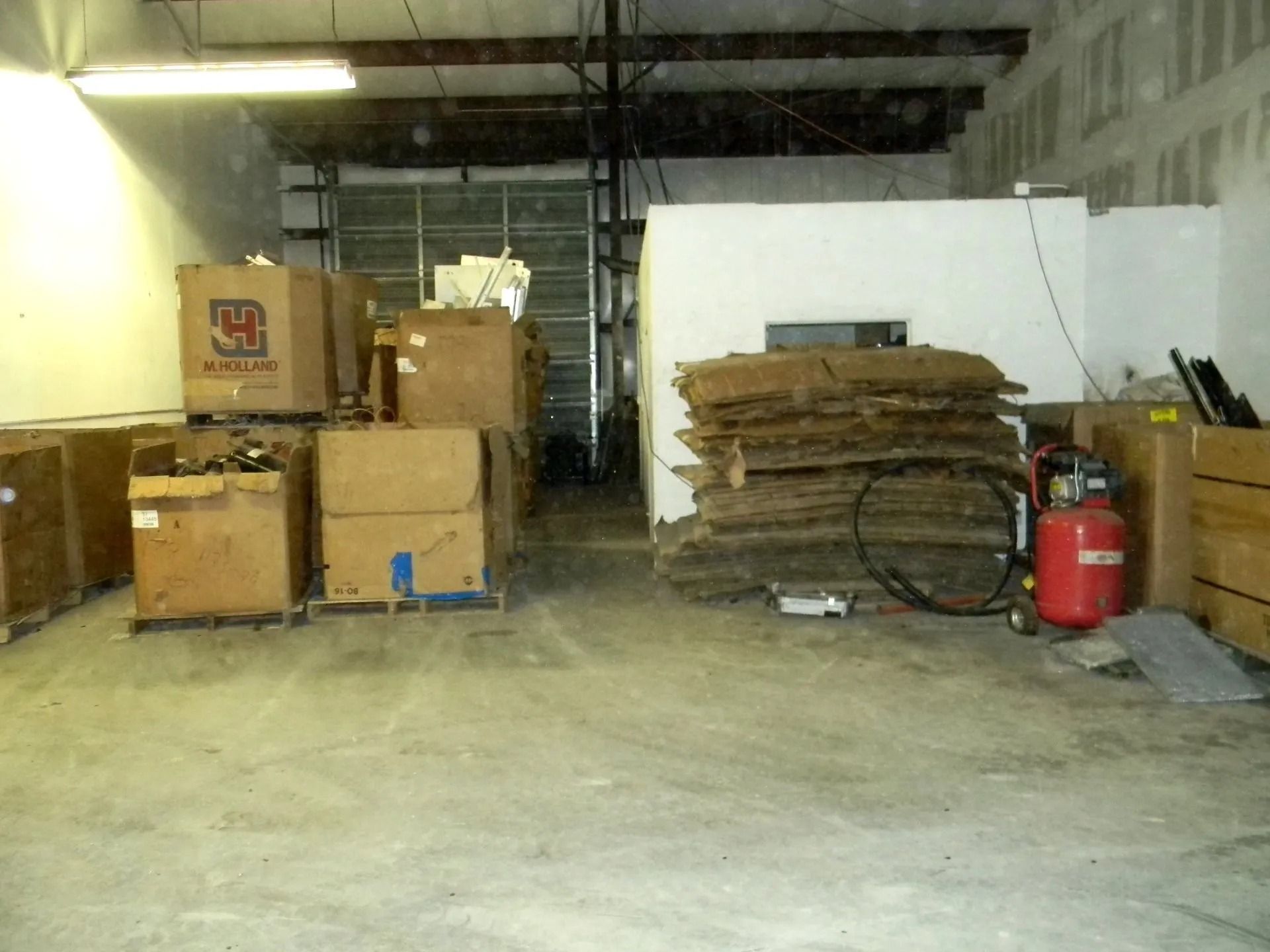 A wide shot of a warehouse interior with cardboard boxes on pallets and a stack of insulation sheets next to a red tank.