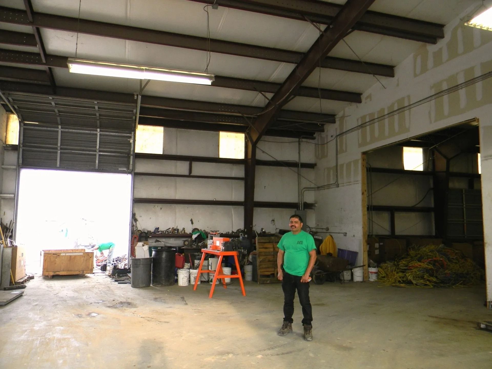 A person in a green shirt stands in a large, industrial warehouse space with an open bay door and scattered equipment.