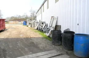 An outdoor area featuring a long white metal wall, industrial waste, scrap metal, and plastic barrels.