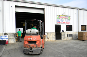 A forklift operator exiting a warehouse bay with a company sign visible on the white metal siding.