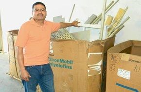A person in an orange shirt and blue jeans leans against a large cardboard bin filled with light-colored scrap materials.