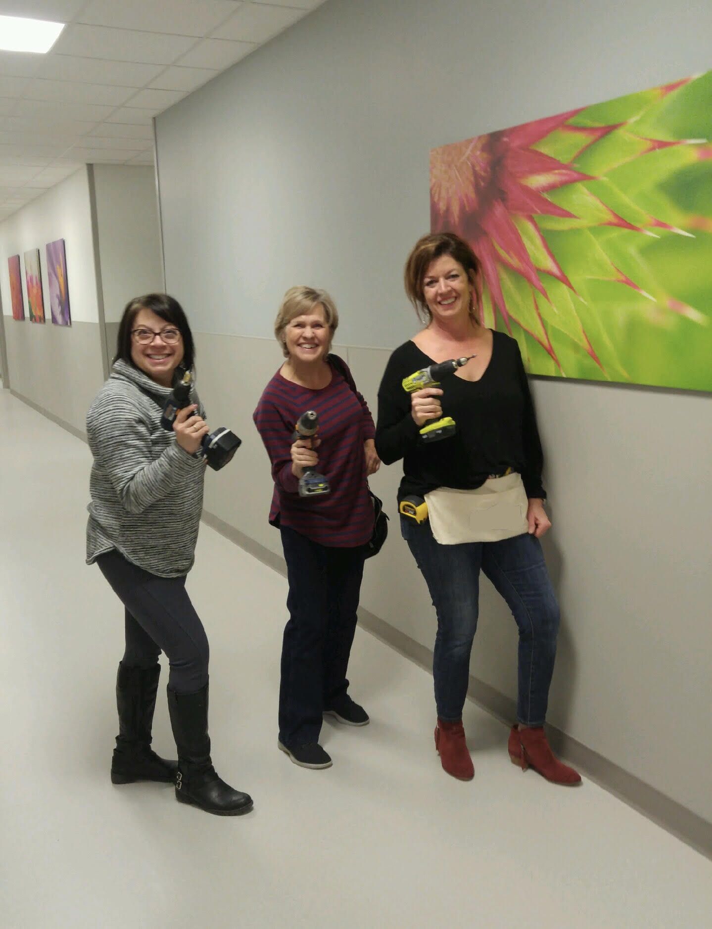 Three women are standing in a hallway with a picture on the wall behind them