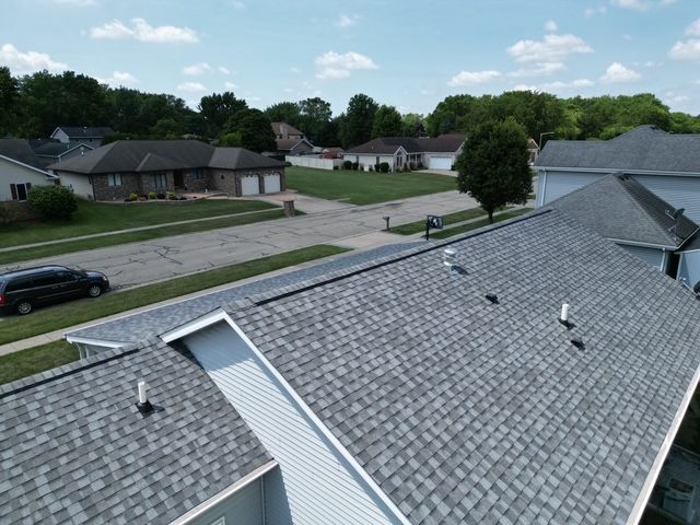 Aerial view of rooftops with gray shingles, neighborhood street, green trees, and blue sky.