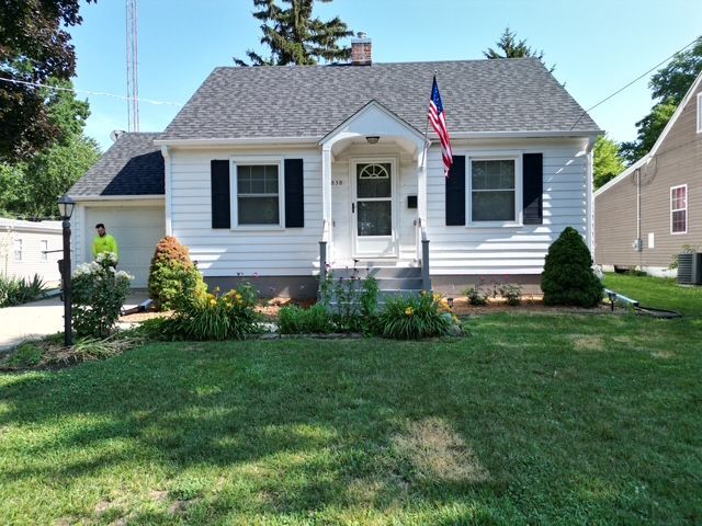 White house with black shutters and an American flag, with a front yard and a person in the yard.