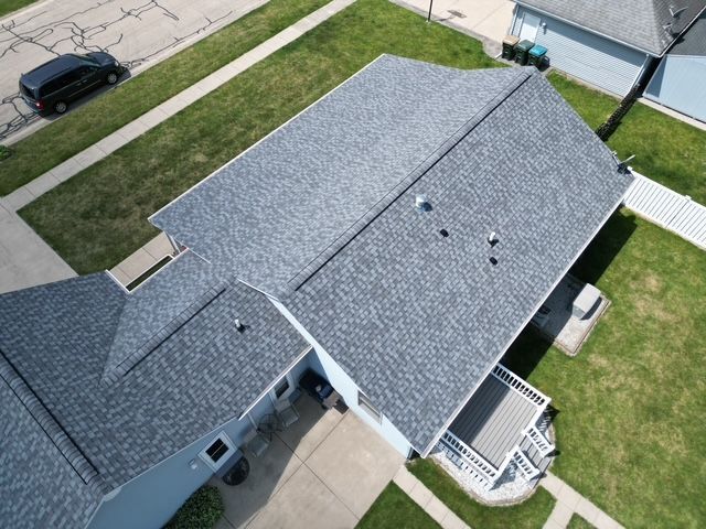 Aerial view of a house with gray asphalt shingle roof, green lawn, and street.