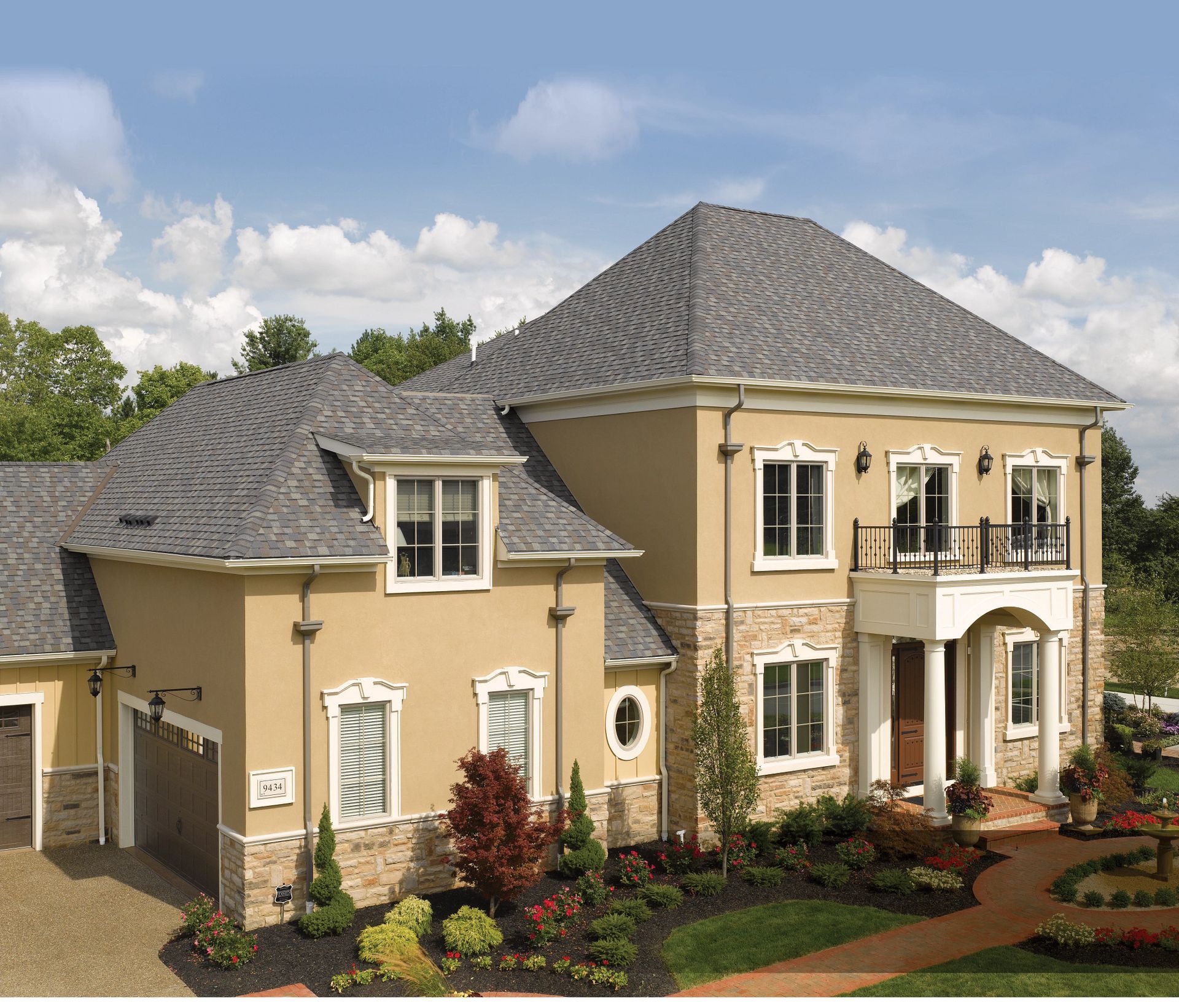 Two-story beige house with gray roof, white trim, columns, and landscaped garden.